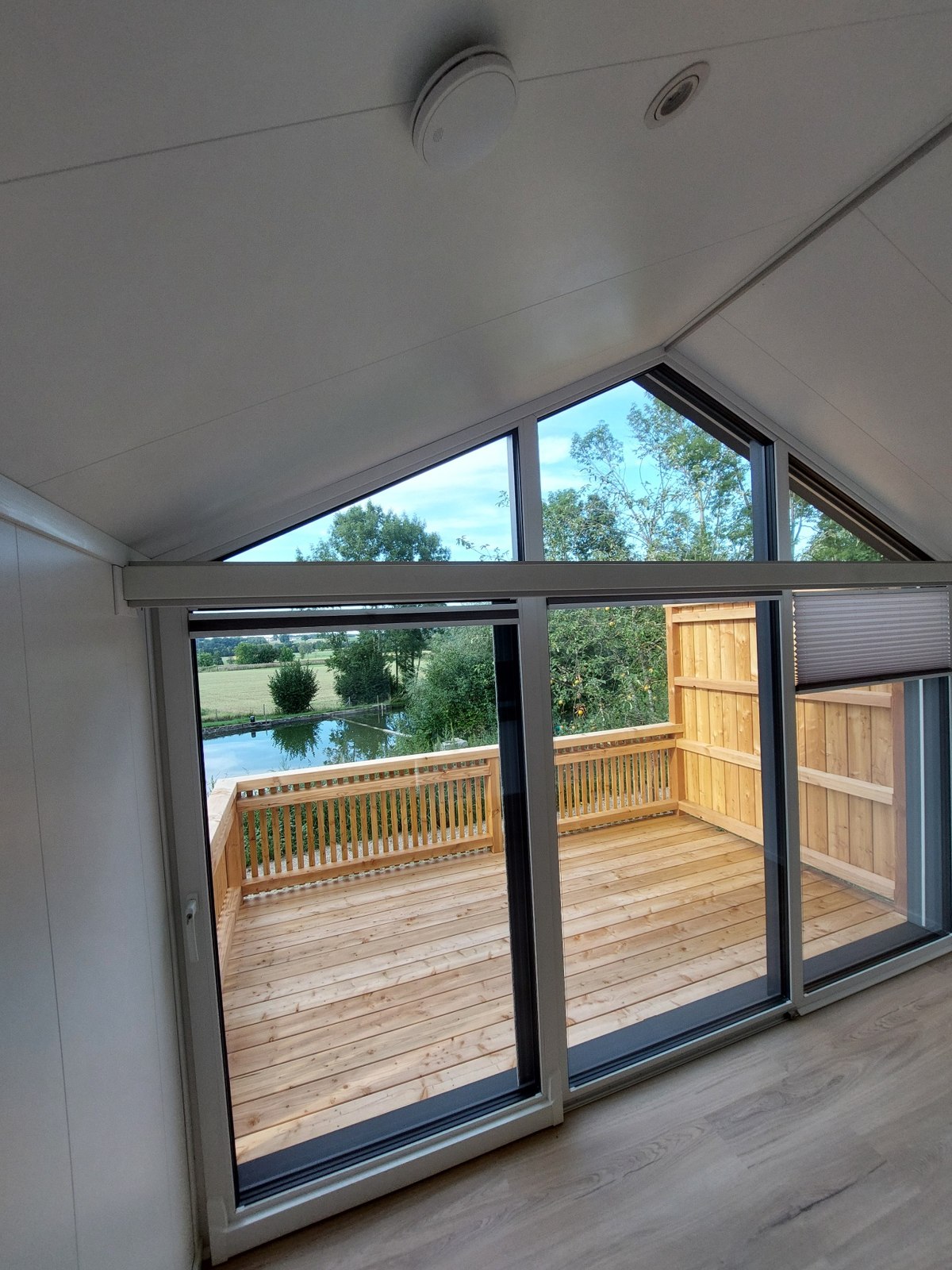 Blick aus dem hellen Wohnraum durch die große Glasfront auf Terrasse, Bungalows und Teich